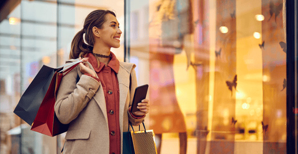 Smiling shopper holding shopping bags and a smartphone while looking into a retail store window.